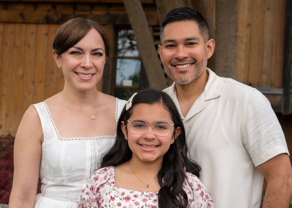 A smiling family of three poses outdoors, featuring a woman in white, a man, and a young girl wearing glasses.
