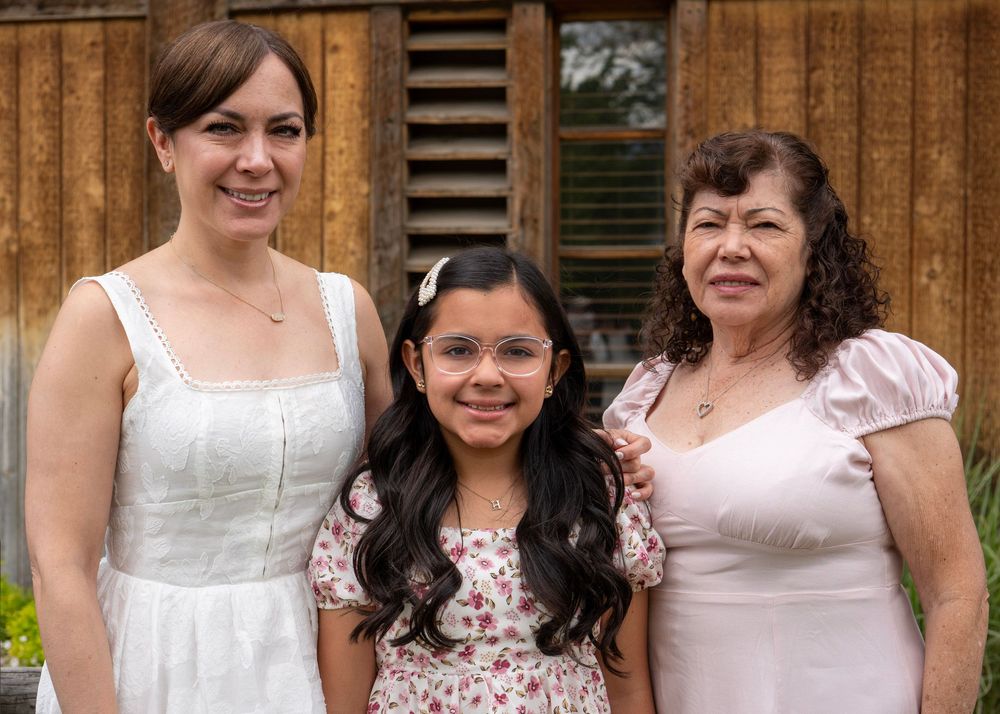 Three generations of women smiling together outdoors, wearing white and floral dresses near a wooden backdrop.
