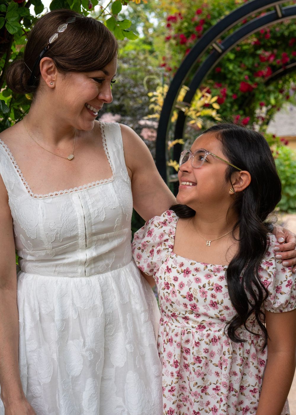 Woman in white dress smiling down at young girl with glasses in a floral dress outdoors.