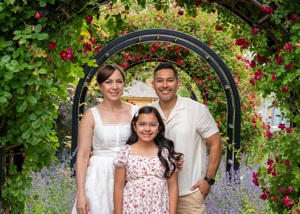 Family of three smiling under a floral arch with red roses and purple flowers in a lush garden.