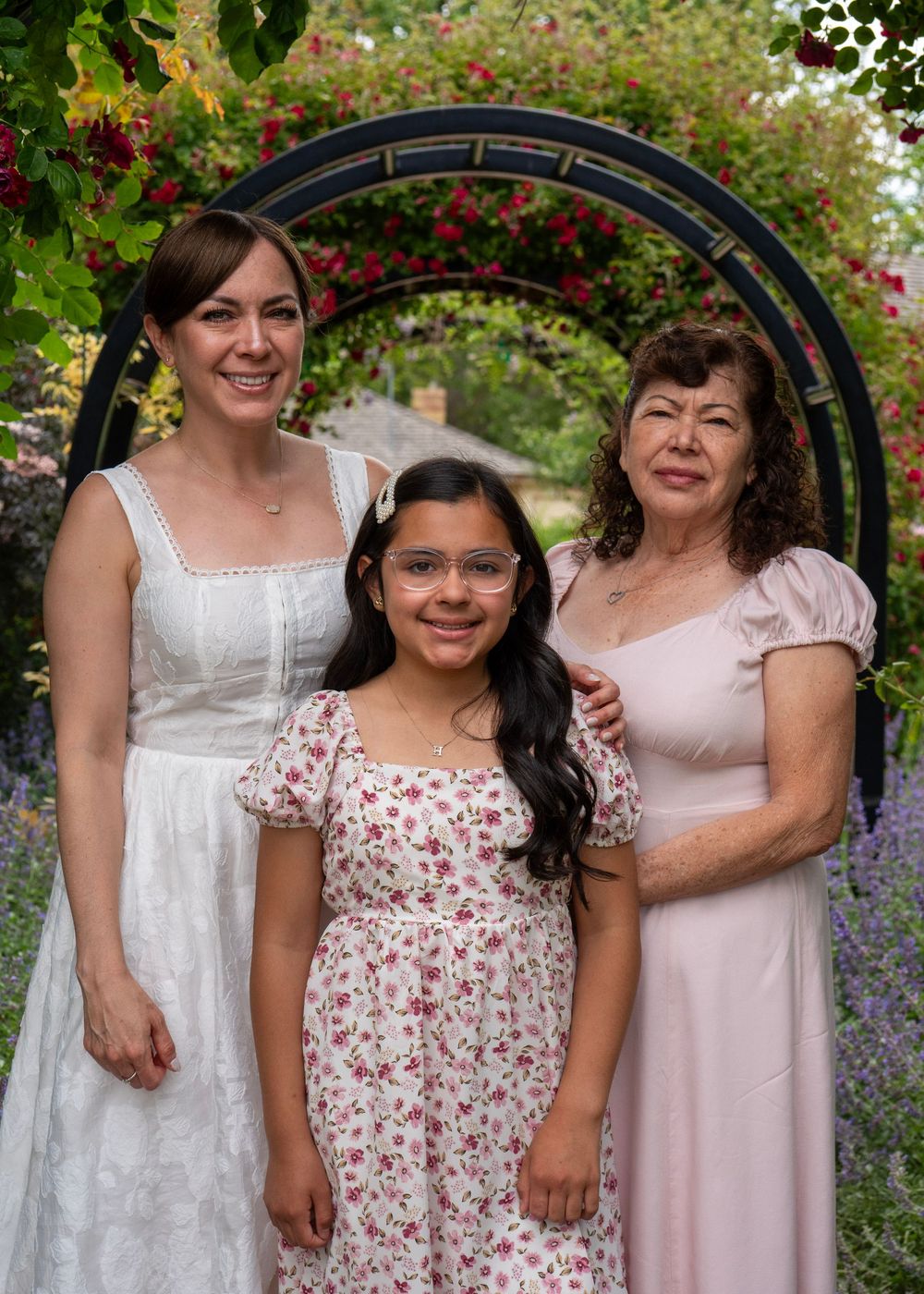 Three generations of women pose in a lush garden with a circular floral arch backdrop.