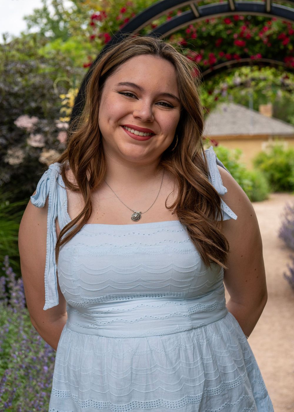 Young woman in white sundress smiles outdoors in a garden with floral arch and lavender blooms.