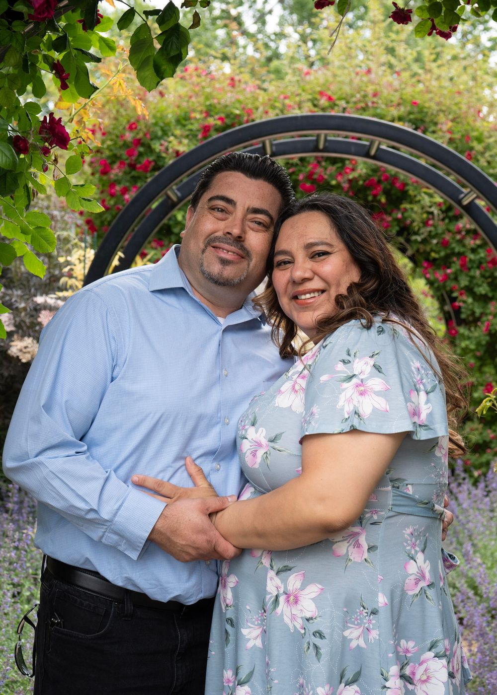Happy couple embracing and smiling under a floral arch with red roses in a beautiful garden.