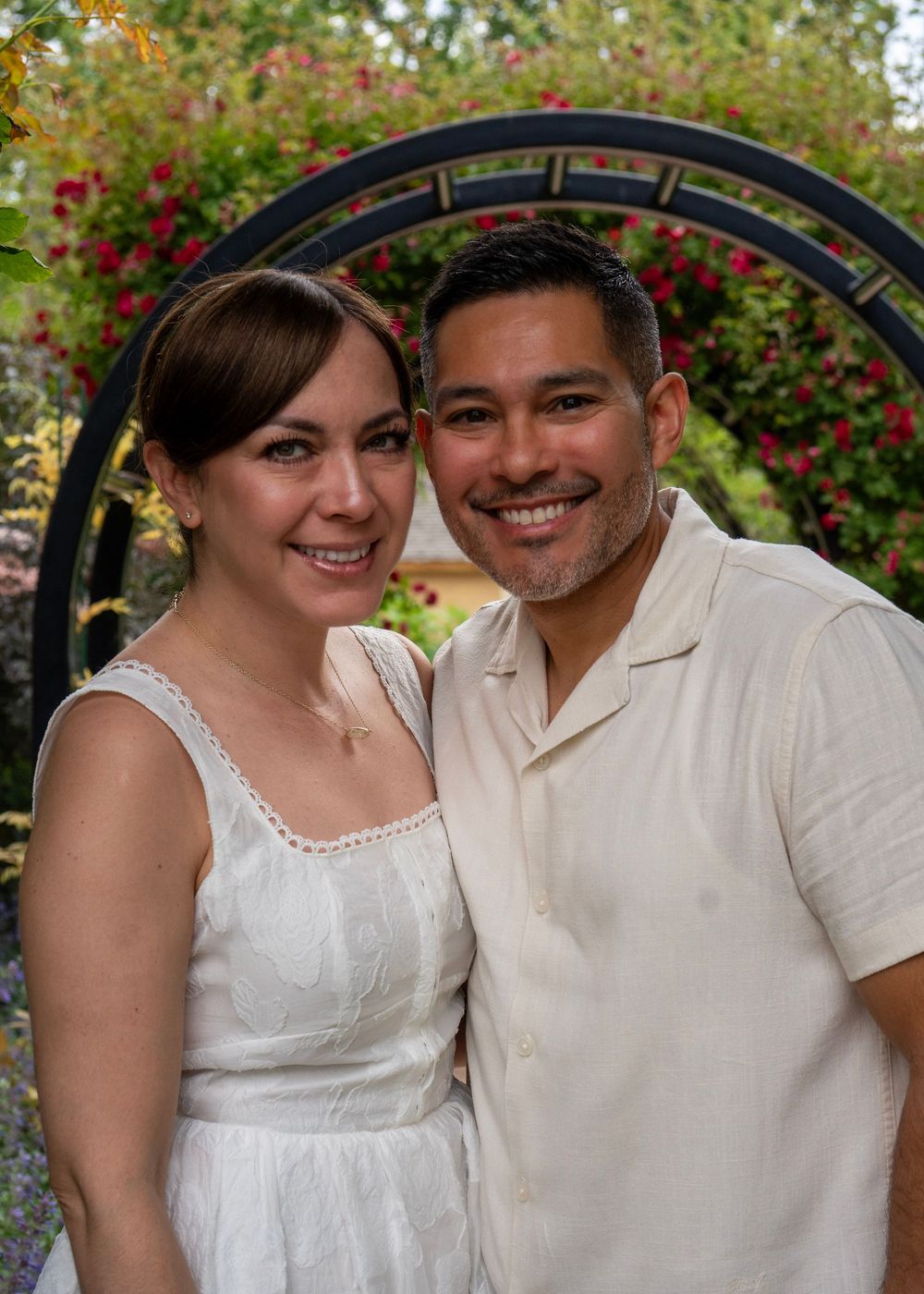 A smiling couple poses together under a floral arch with red roses in a garden setting.