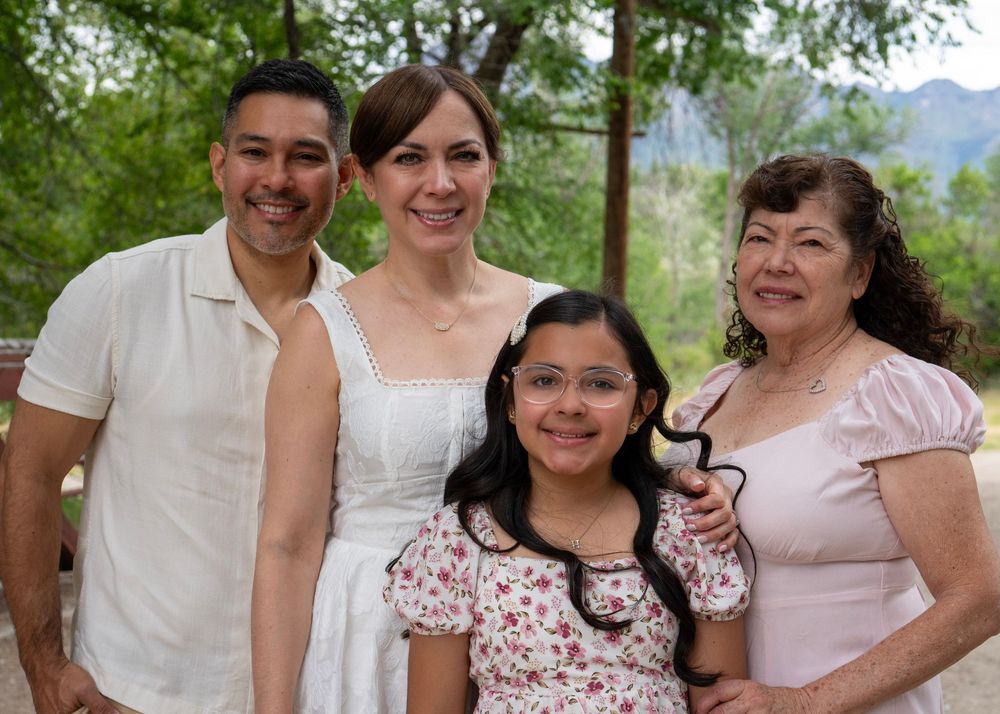 Family of four smiling outdoors with mountains behind them on a sunny day.