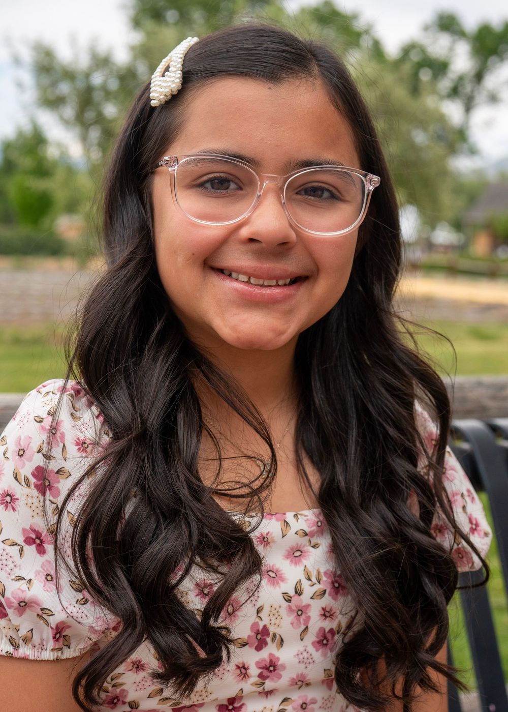 Young girl with long dark hair, clear glasses, and floral top smiling outdoors.