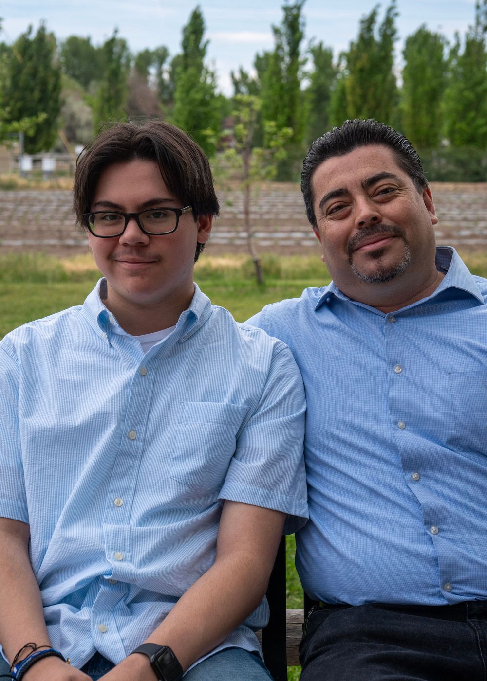 Two men in light blue shirts smile while sitting outdoors with trees in the background.