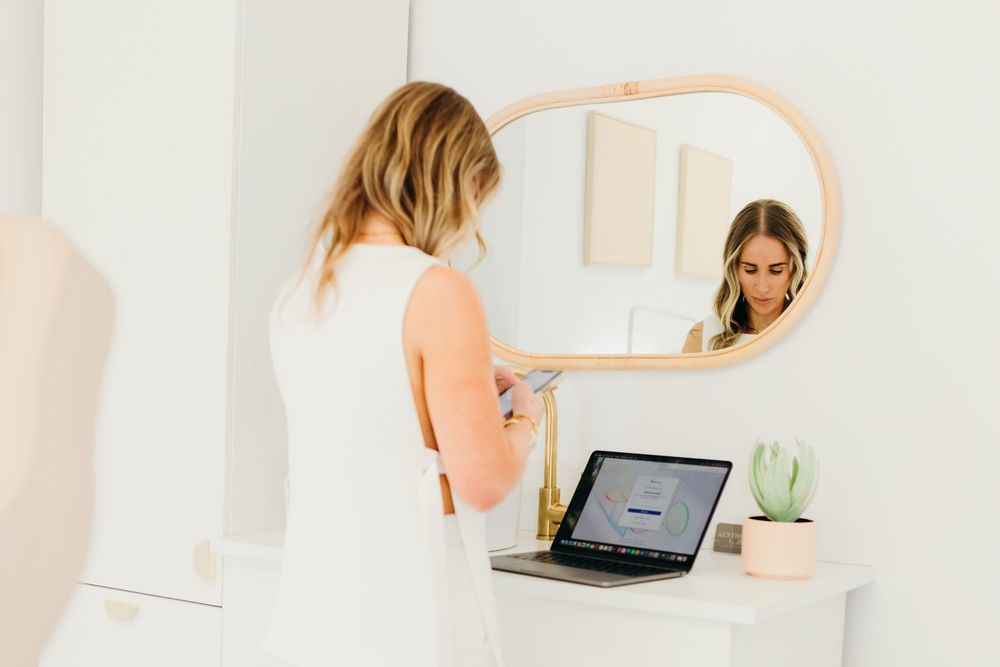 Blonde woman checking phone reflected in arched mirror above white desk with laptop.