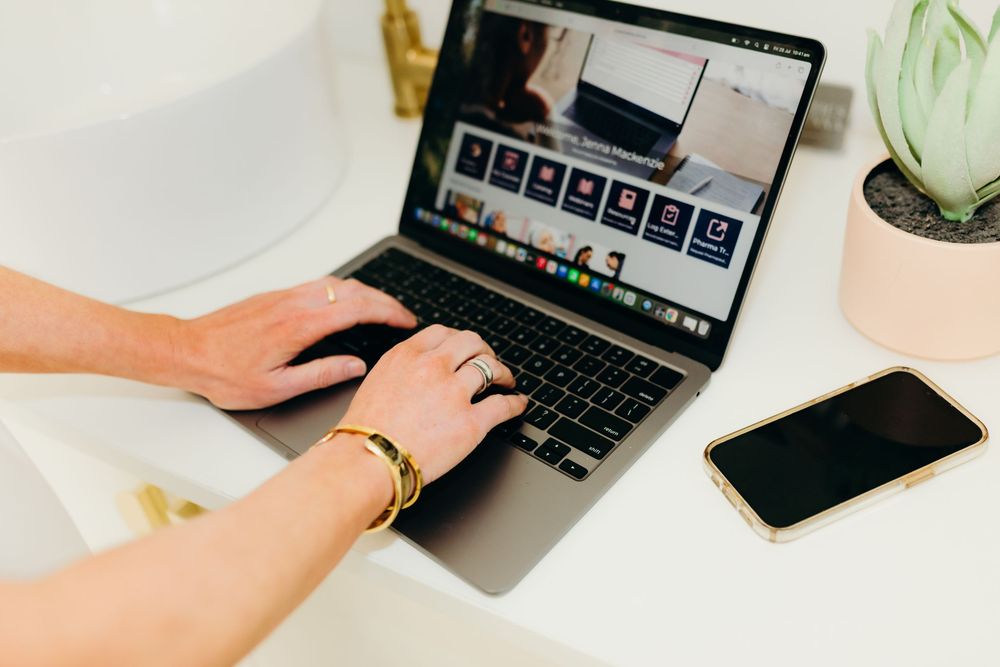 Close-up of hands with gold bracelet typing on MacBook laptop beside smartphone.