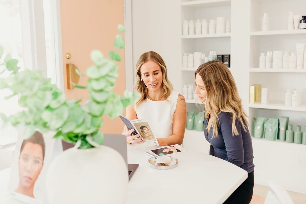 Two women consulting over skincare magazines at white table in elegant beauty studio.