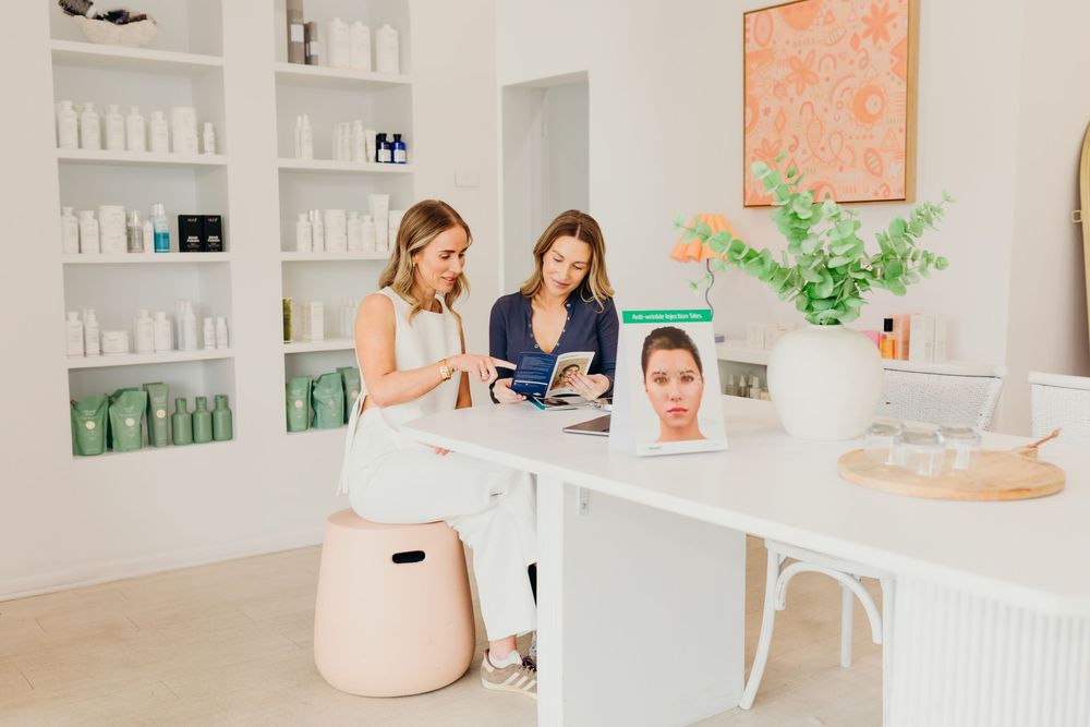 Two women consult at a bright skincare clinic reception desk with product shelves and a facial analysis screen.