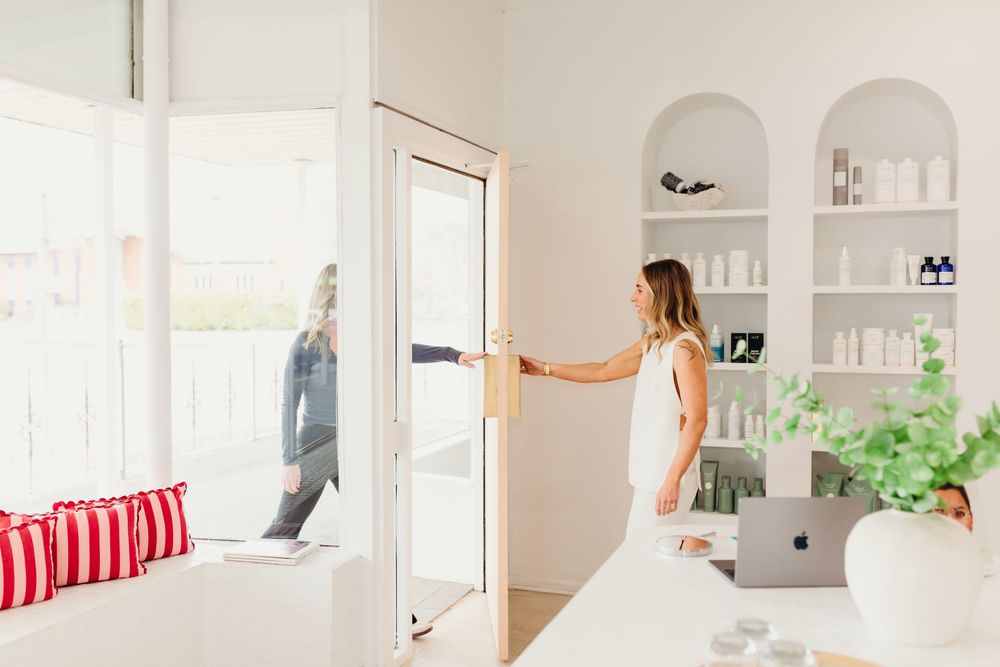 A woman in white opens the door of a bright, minimalist skincare clinic with product-lined shelves.