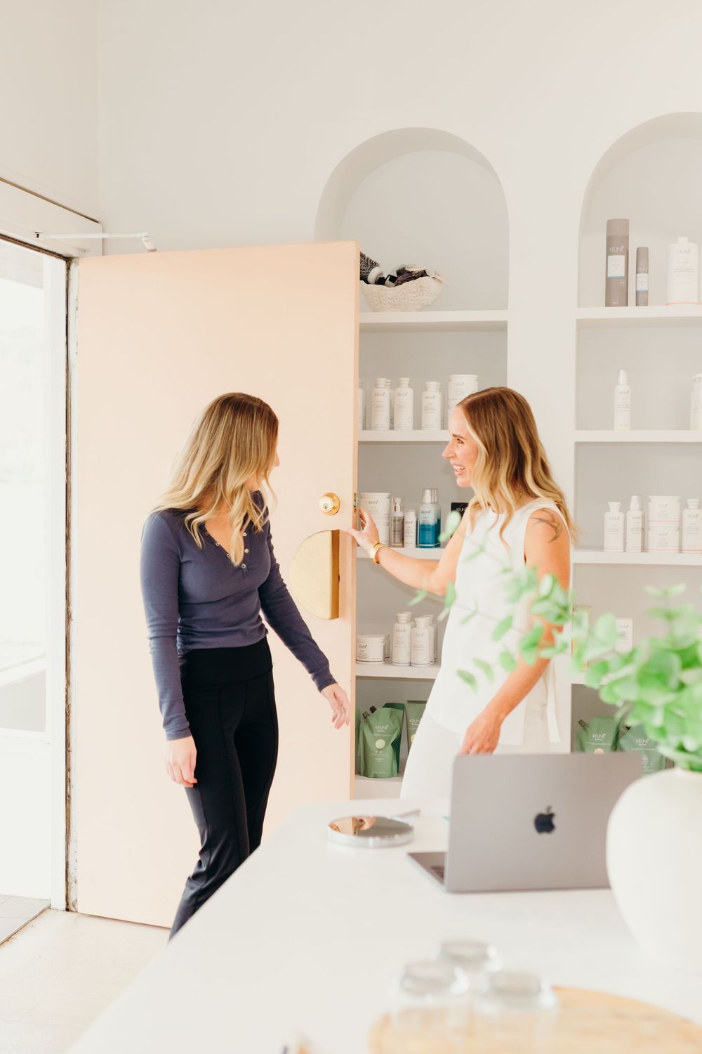 Two women chat near arched shelves of skincare products in a bright, modern beauty clinic interior.