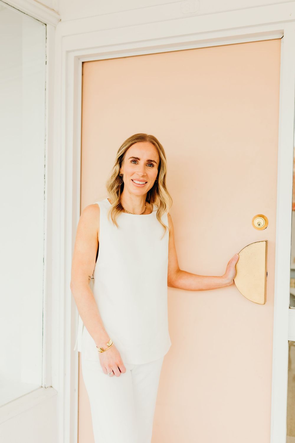 A smiling blonde woman in white leans against a peach door with a gold handle in a bright space.