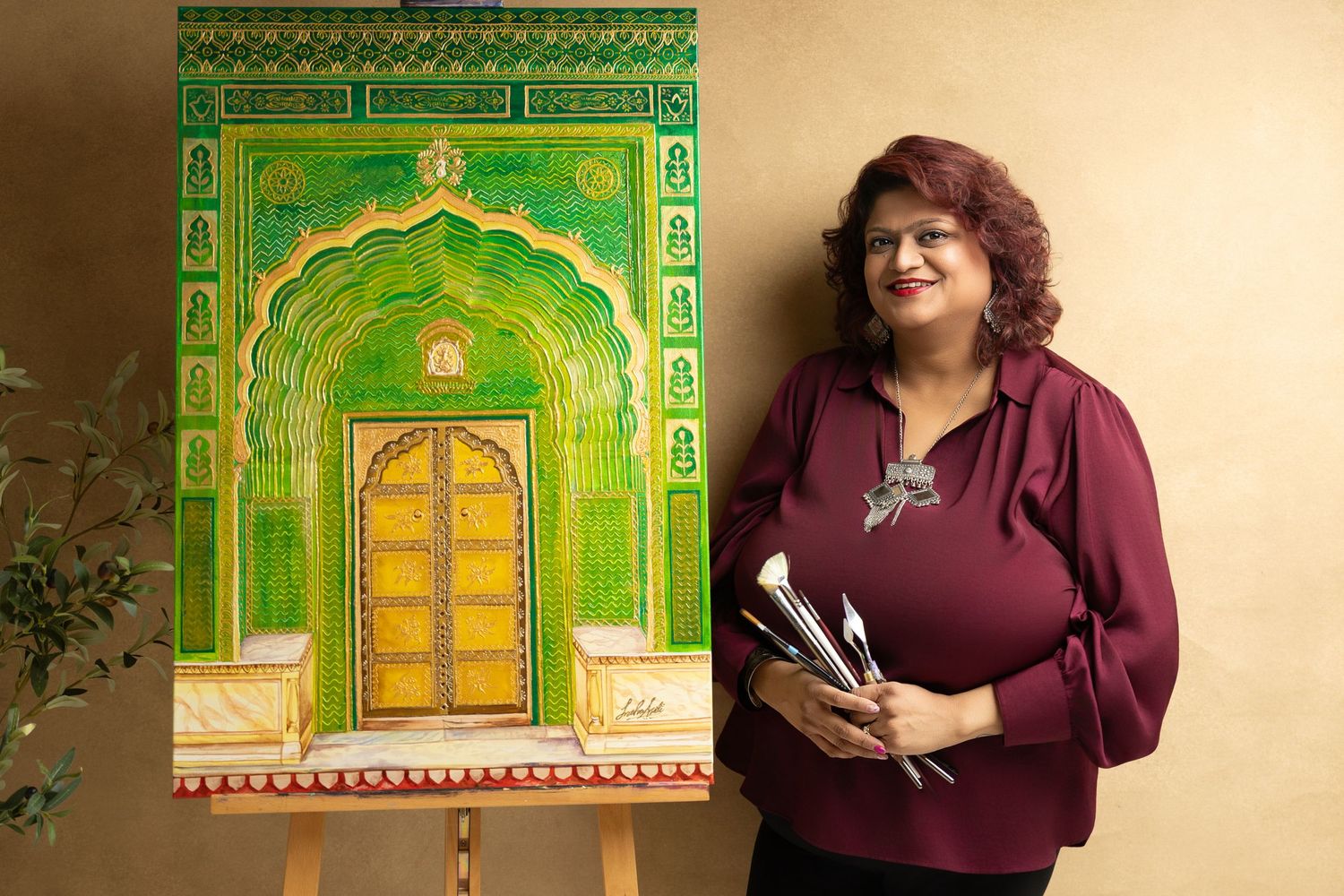 Woman holding paintbrushes stands beside a vibrant painting of an ornate green and gold Indian archway on an easel.