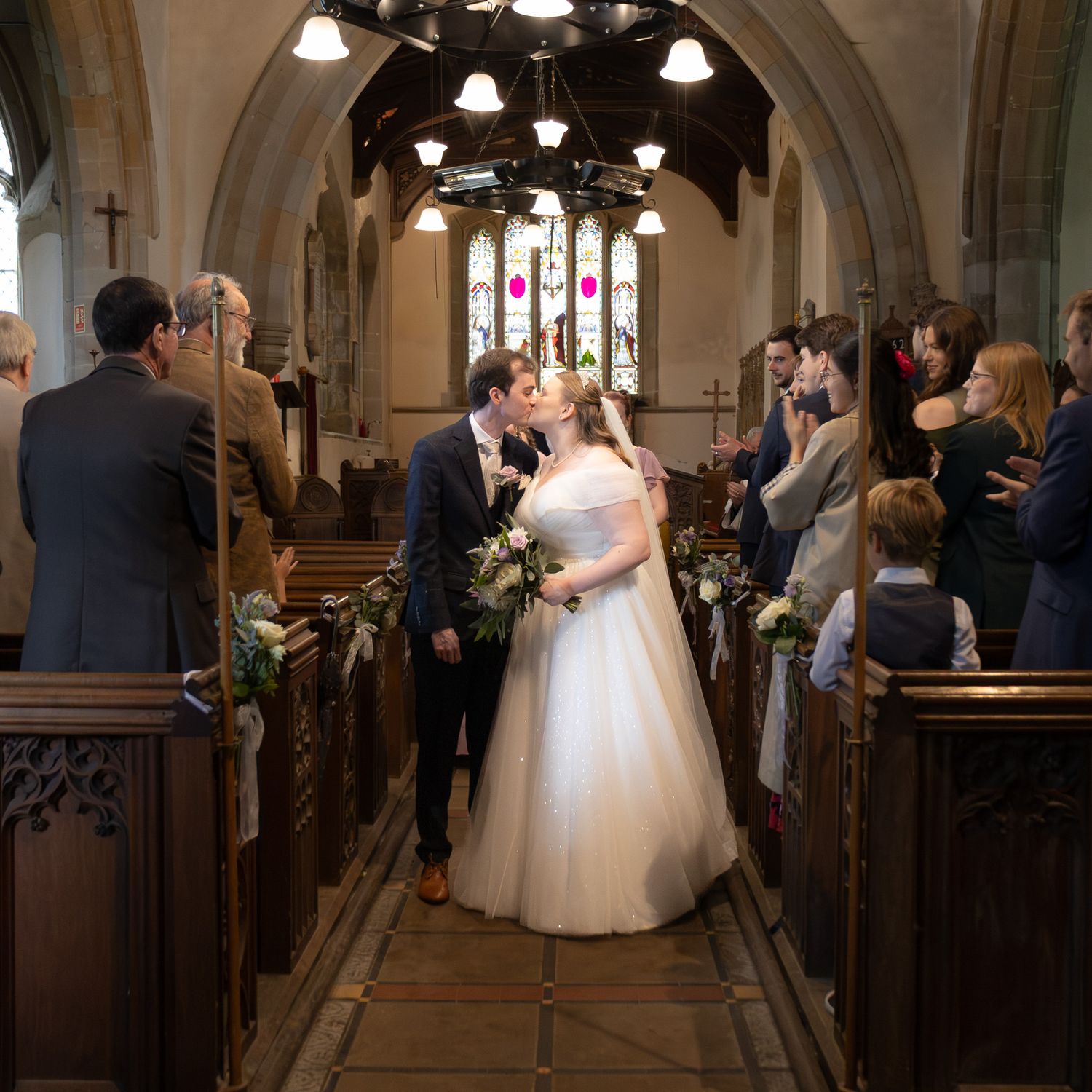 Bride and groom walk hand in hand down the aisle, smiling, as guests applaud in an elegant venue.