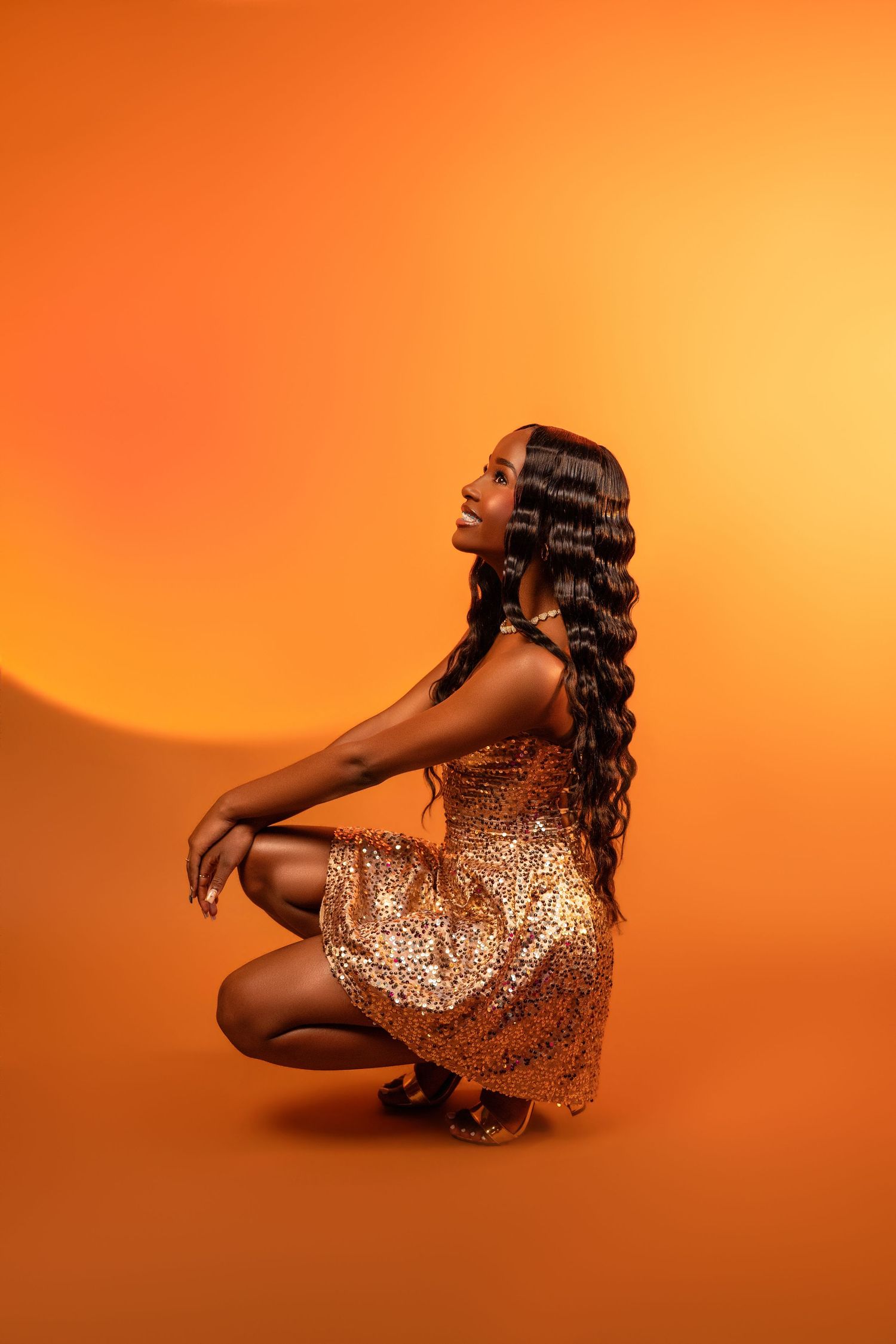 Black woman in gold sequin dress crouching gracefully against vibrant orange background with long wavy hair.