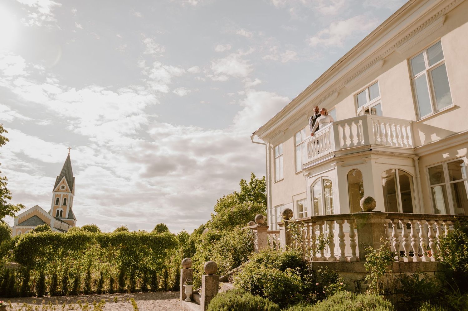 Bride standing on white balcony of elegant villa with church steeple and lush greenery in background.