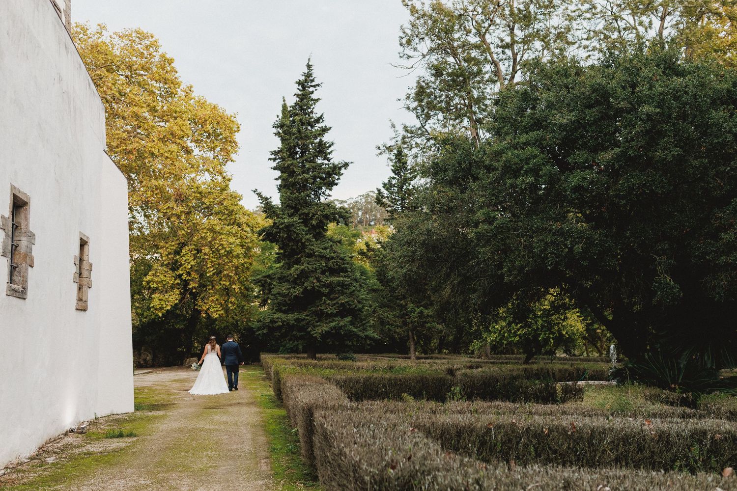 Distant couple in white walking along a stone wall pathway beside a church building and autumn trees.