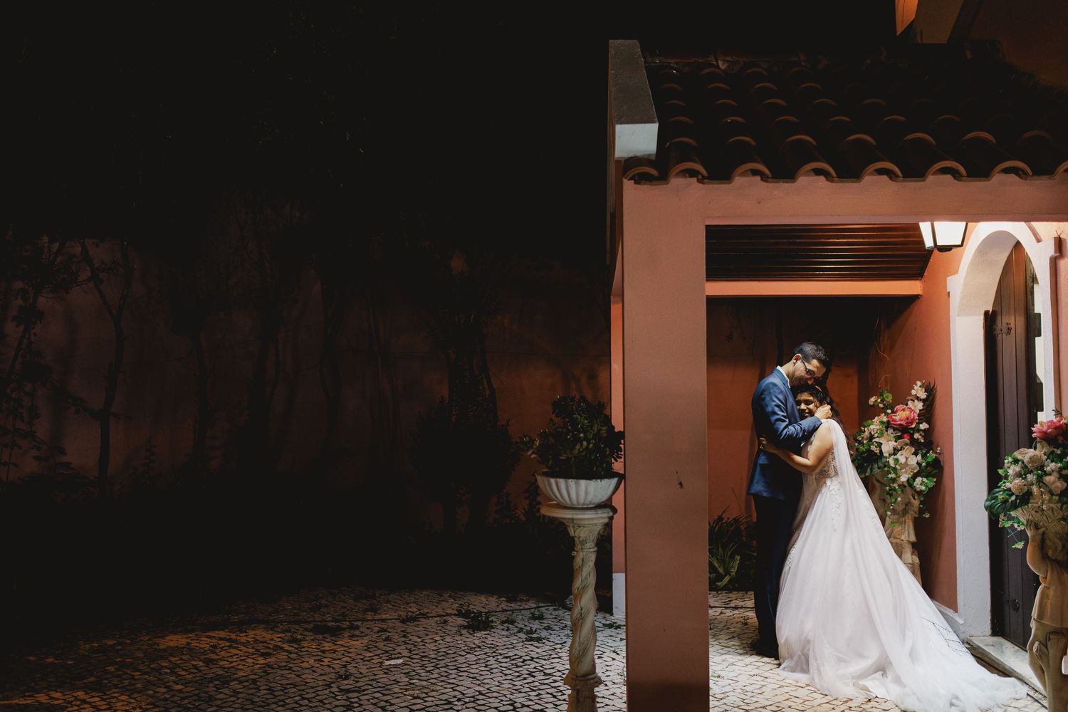 A couple embraces under a Mediterranean-style terracotta archway surrounded by lush garden scenery at dusk.