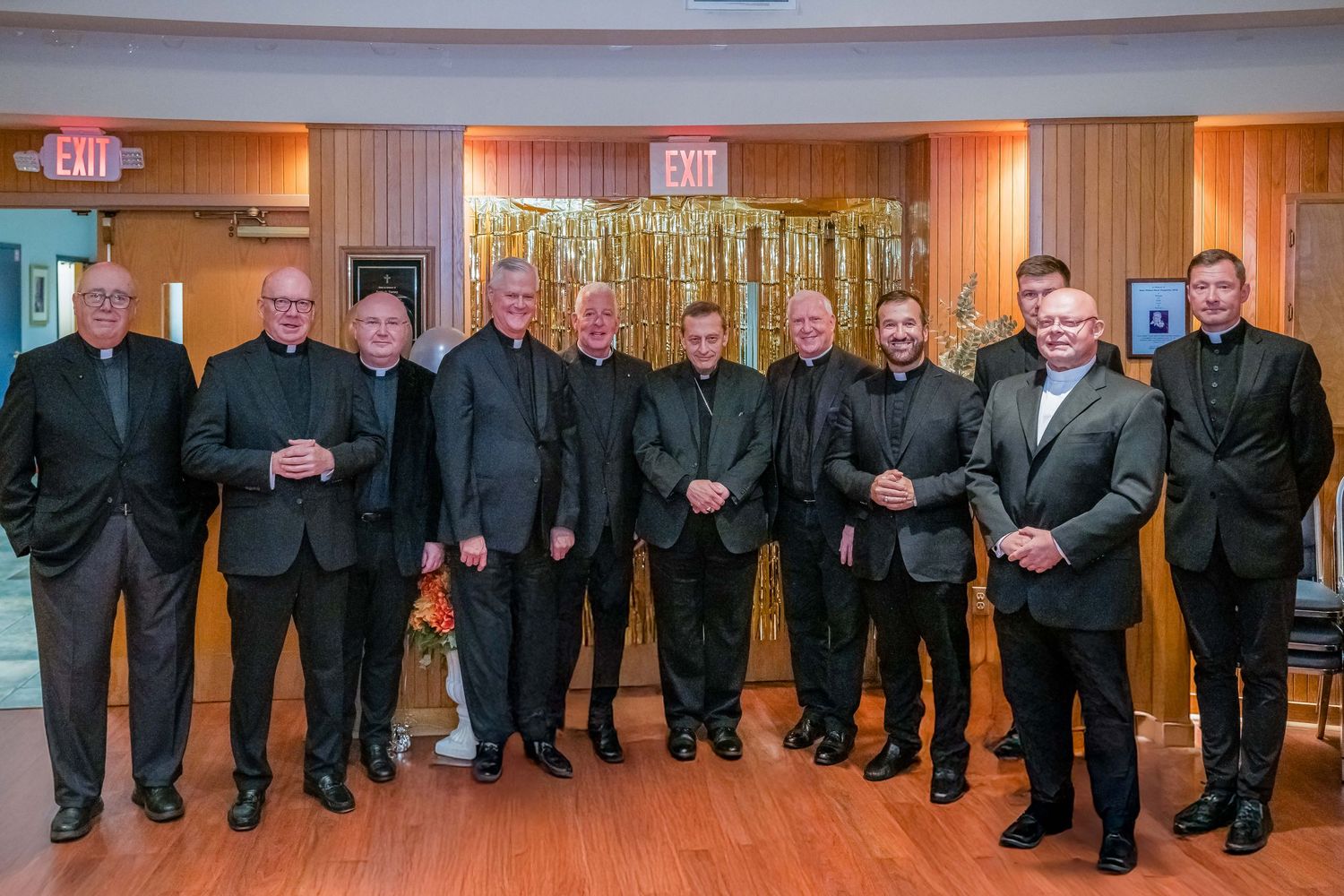 Group of men in formal black attire posing together in front of gold tinsel backdrop at an elegant event.