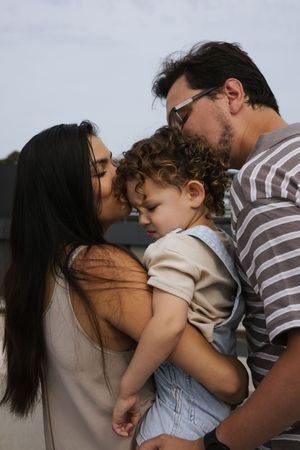 Loving parents kissing their curly-haired toddler on the cheeks during an outdoor family photo.