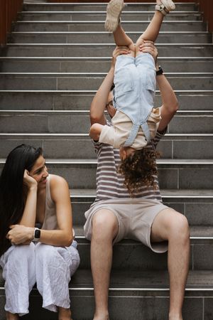 Father playfully holding toddler upside down on steps while mother watches and smiles.