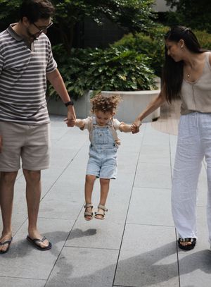 Parents swinging their curly-haired toddler in denim overalls between them on a sunny plaza.
