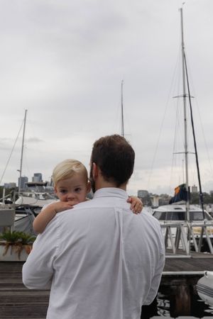 Father holds blonde toddler at a marina with sailboats in the background.