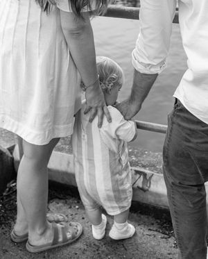 Black and white photo of parents holding their toddlers hands from behind near a waterfront.