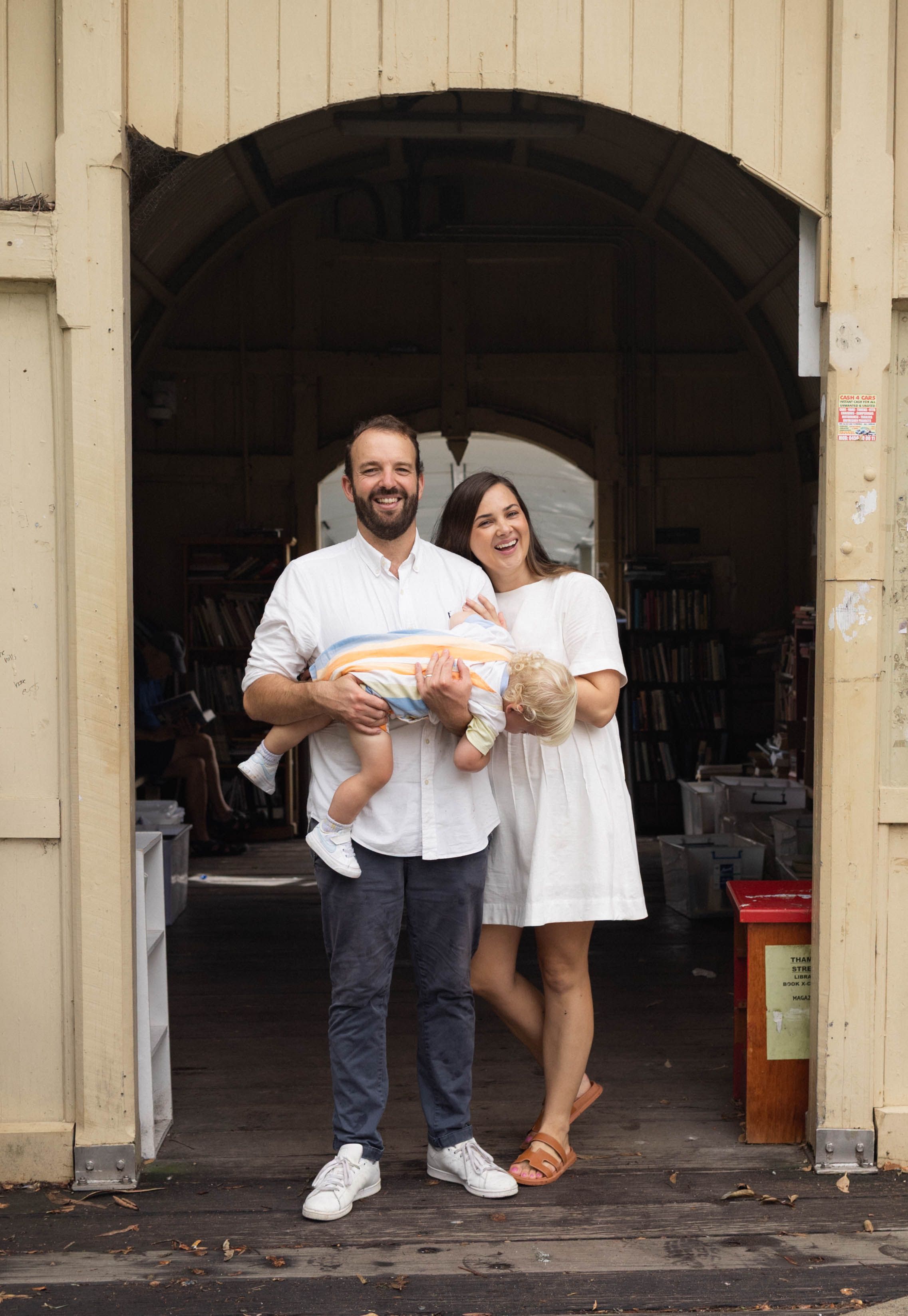 Smiling couple stands in arched doorway of a bookshop holding their baby.