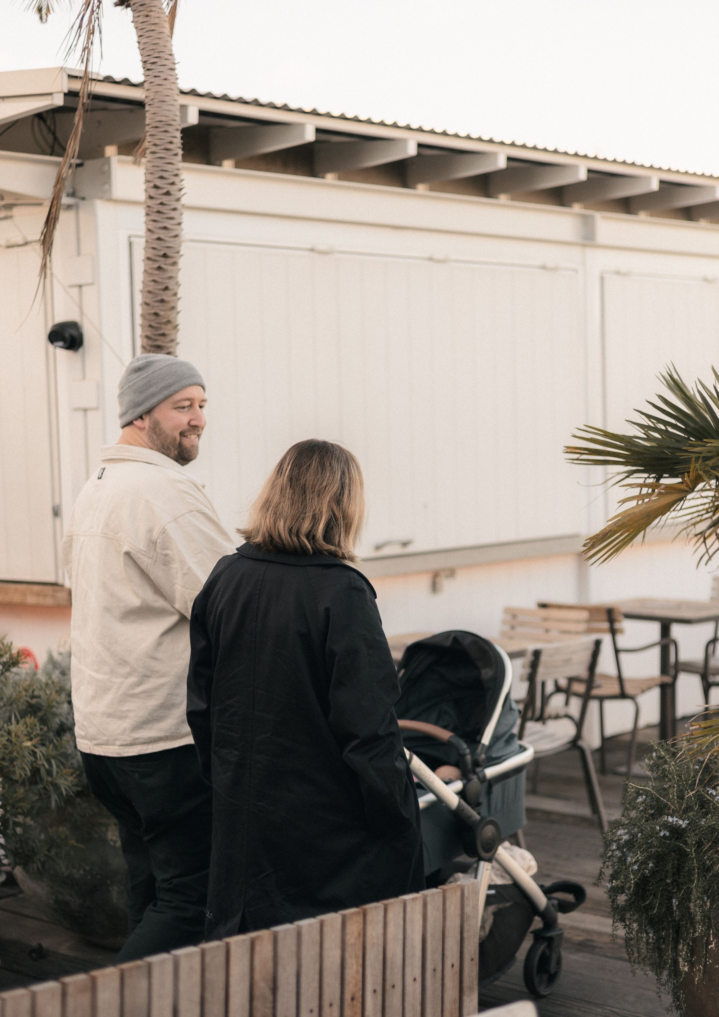 Couple with a pram chat outside a cafe near palm trees at dusk.