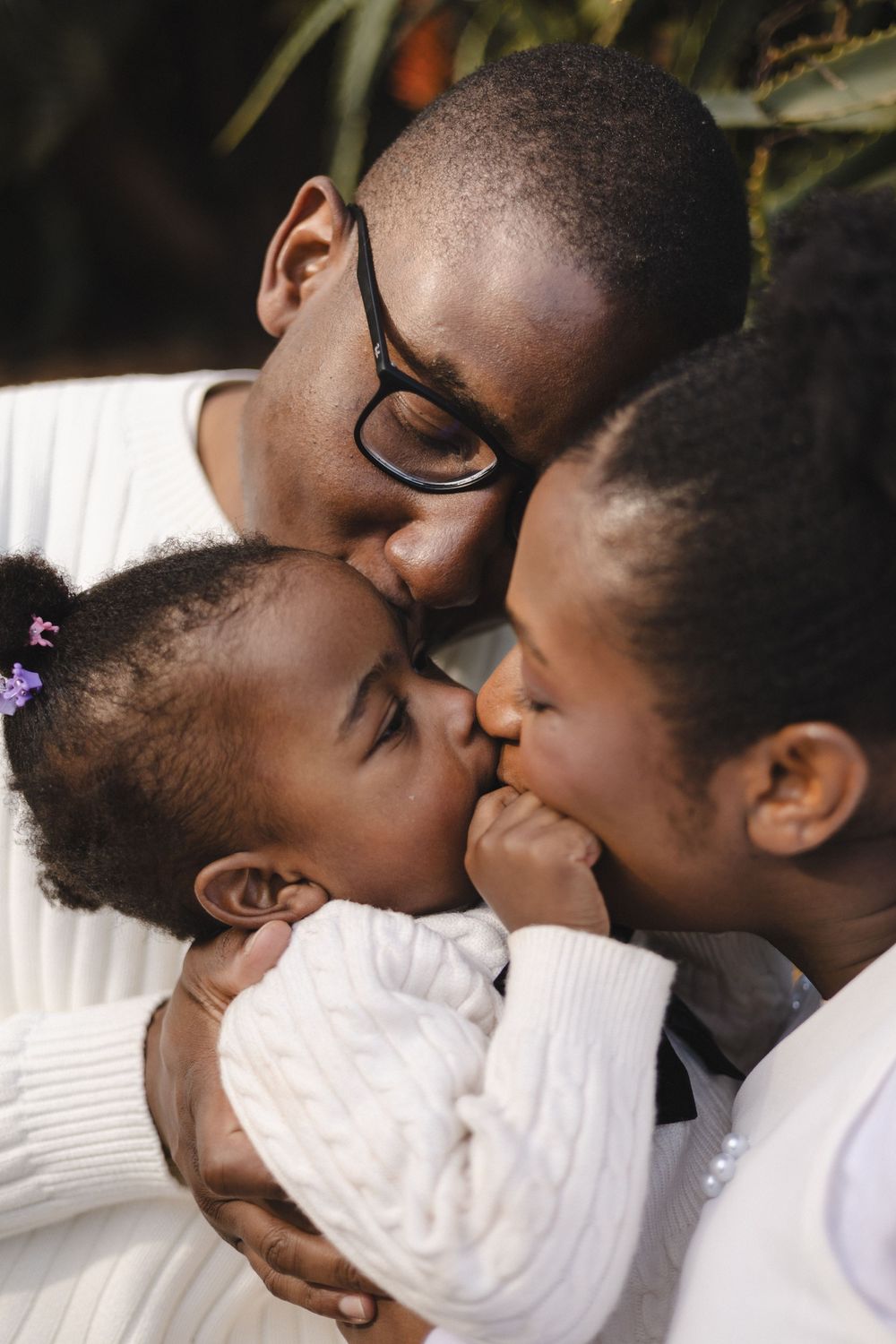 A father with glasses shares a tender moment kissing his two young daughters, all dressed in white sweaters.