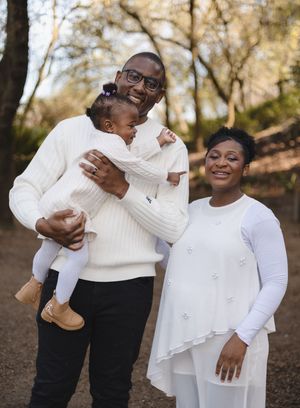 Happy family in white outfits posing outdoors, father holding baby girl, mother smiling beside them in wooded setting.