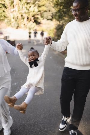 Happy Black family swinging toddler girl in white dress outdoors on a sunny day.