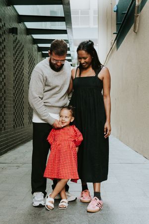 Smiling interracial family of three posing in an urban alleyway, daughter wearing red gingham dress.