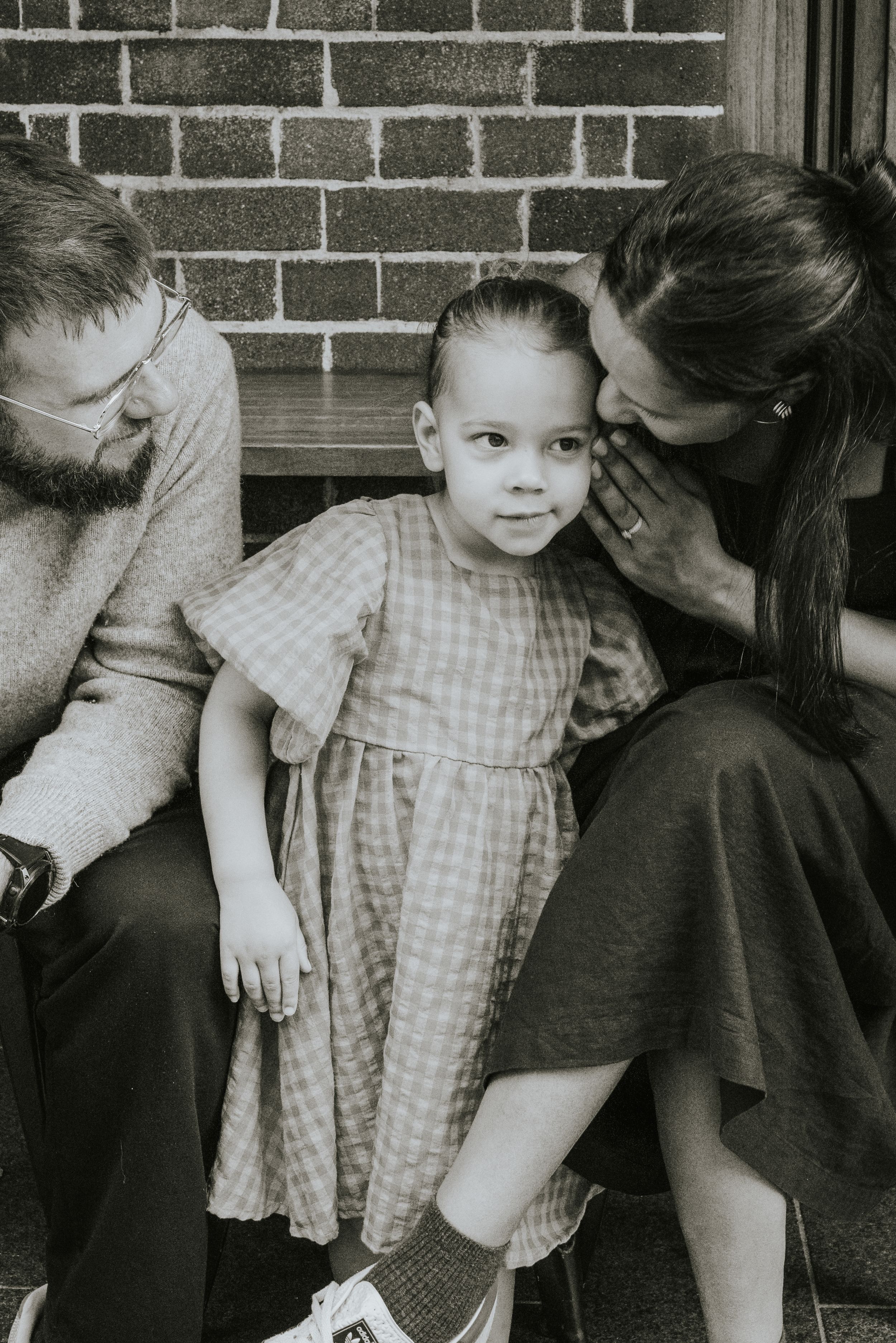 Black and white photo of a young girl in a plaid dress sitting between her parents on steps near a brick wall.