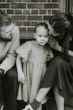 Black and white photo of a young girl in a plaid dress sitting between her parents on steps near a brick wall.