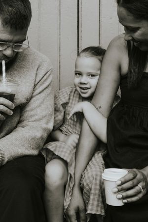 Black and white photo of parents and young daughter sitting together sharing drinks outdoors.