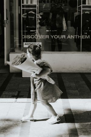 Black and white photo of a young girl in a dress carrying a large bag outside a store reading Discover Your Anthem.