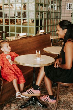 A woman and young girl in a red dress sit at a cafe table, smiling, woman wearing Adidas sneakers.