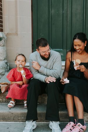 A bearded man and woman laugh while eating ice cream on steps with a young girl in a red dress.
