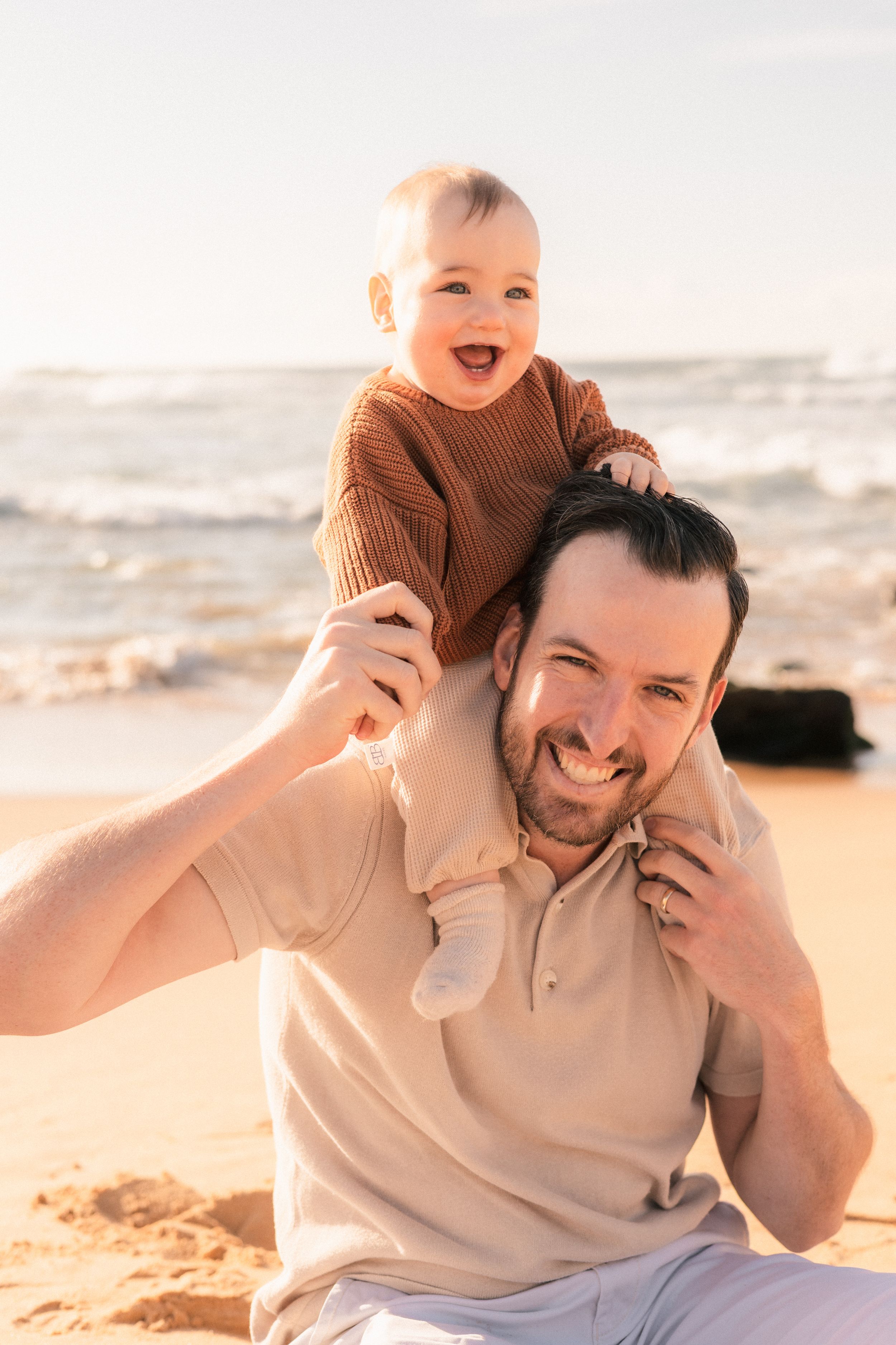 Smiling father holds laughing baby on his shoulders on a sunny beach.