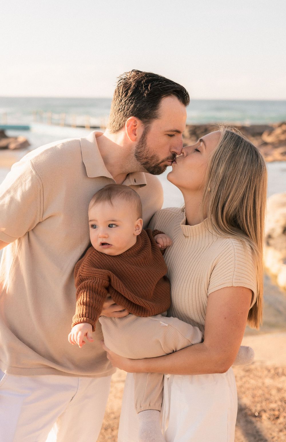 Happy young couple kissing at the beach while holding their baby in a brown sweater.