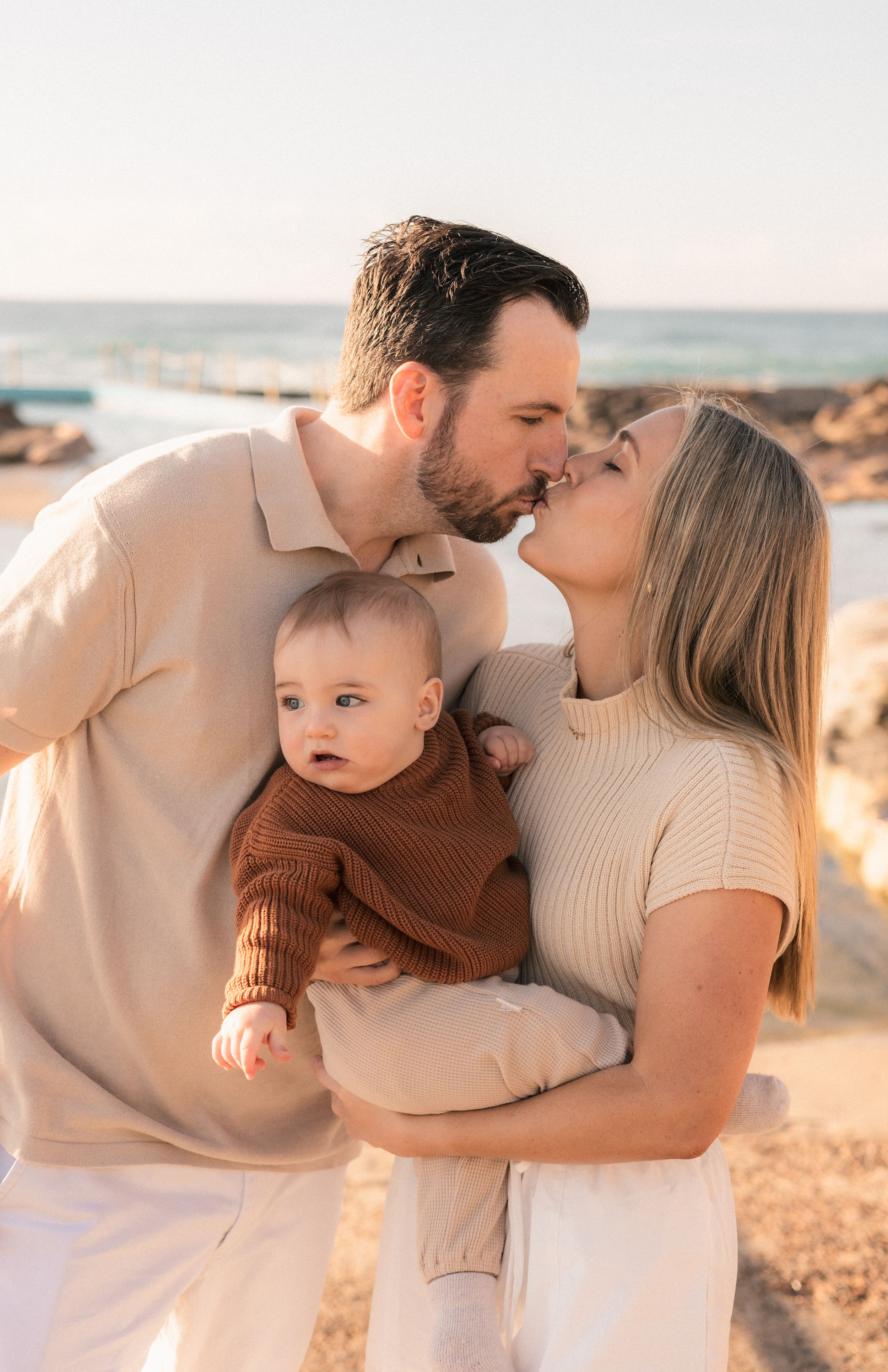 Happy young couple kissing at the beach while holding their baby in a brown sweater.