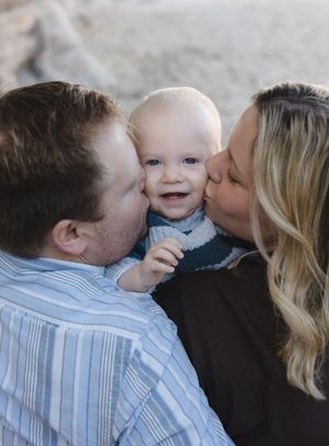 Two parents lovingly kiss their smiling baby on the cheeks in a sweet outdoor family portrait.