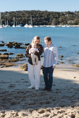 Family with baby walking on sandy beach with sailboats and blue water in background on sunny day.
