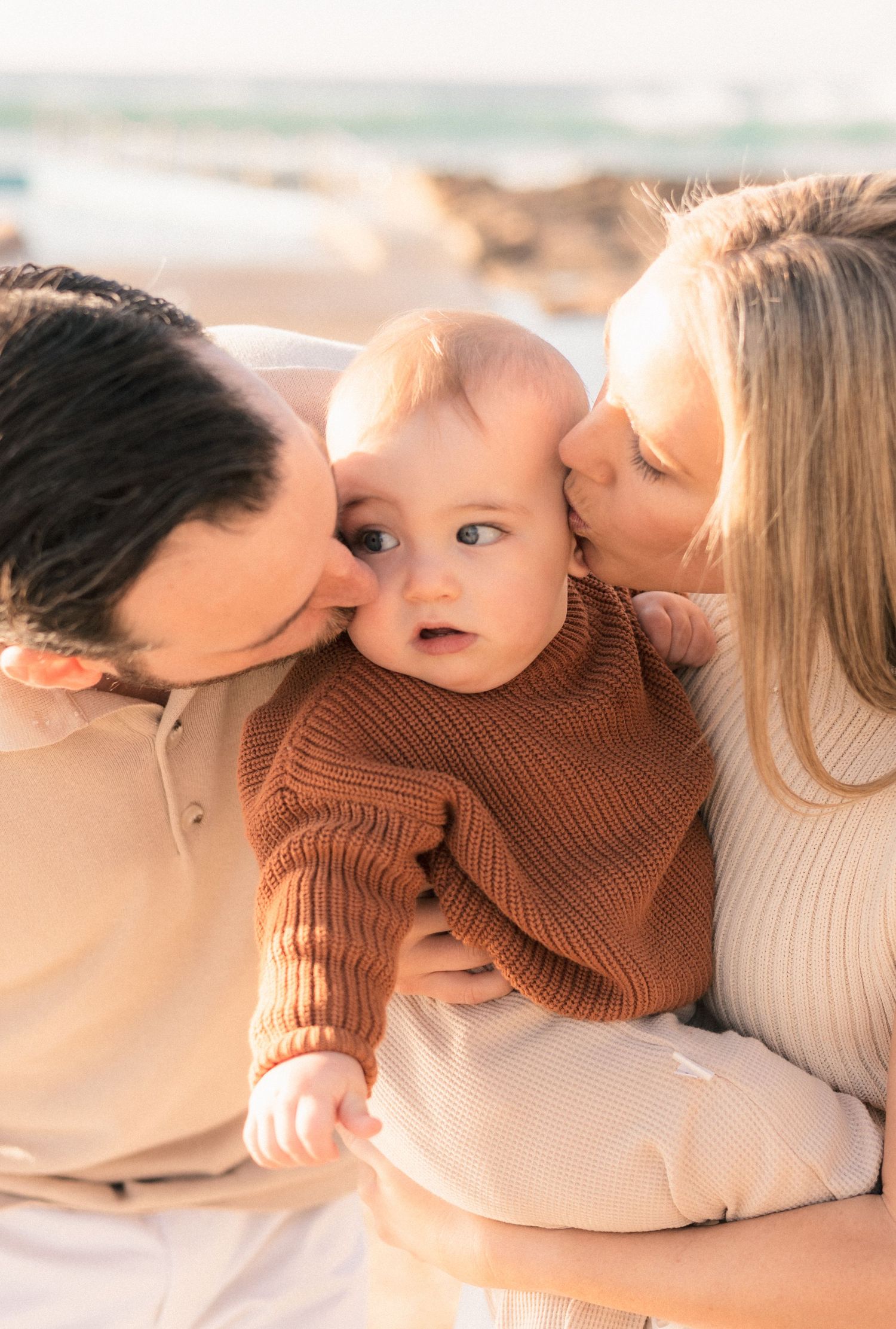 Parents kissing their baby on the cheeks at a sunny beach, baby in brown knit sweater looks surprised.