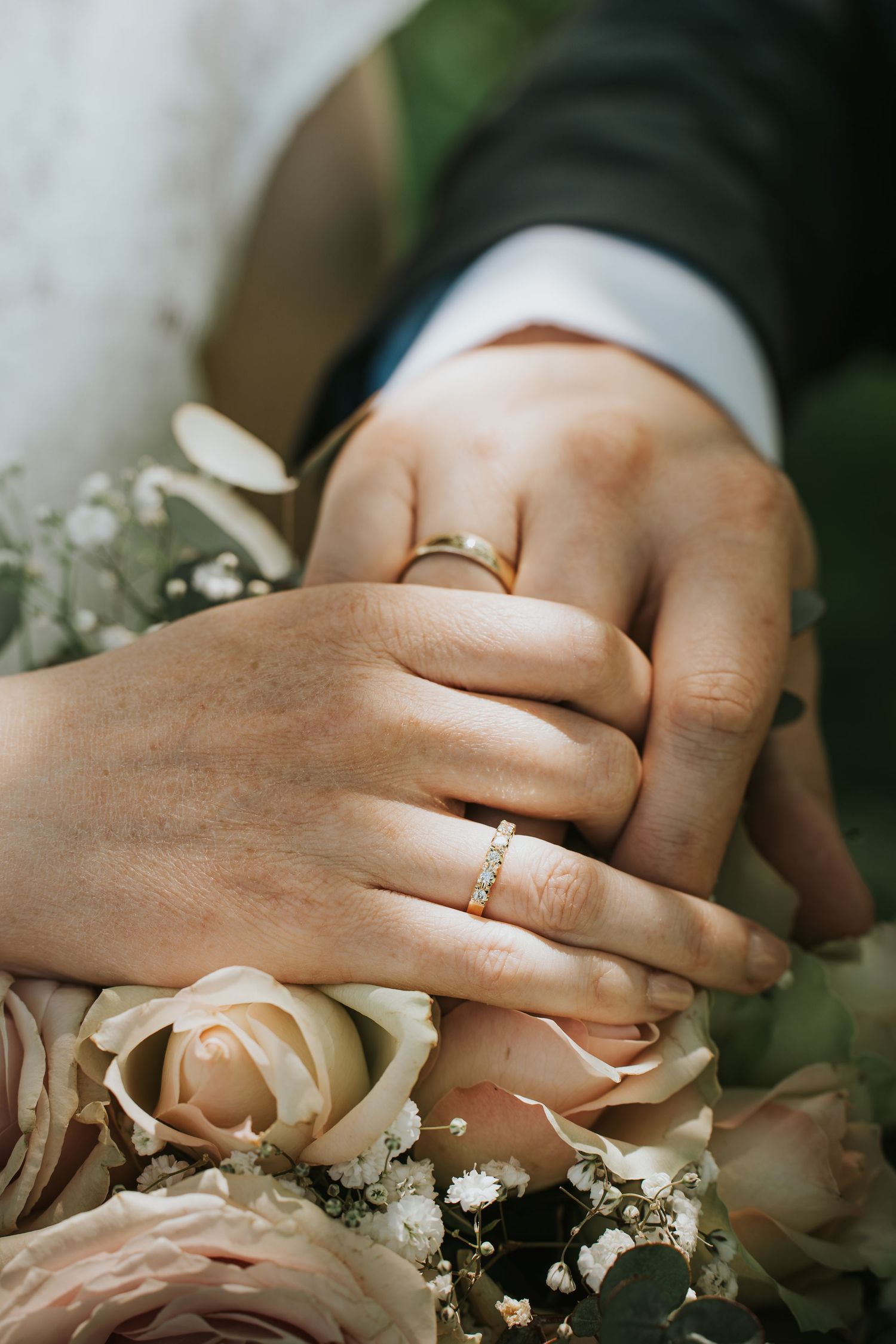 Newlywed couple displaying gold wedding rings on hands resting over a bouquet of blush pink roses.