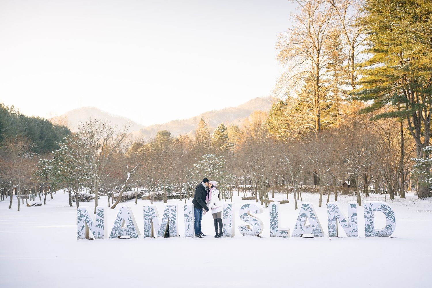 Couple embracing among large NAMILAND letters in a snowy winter landscape with mountains and trees at golden hour.